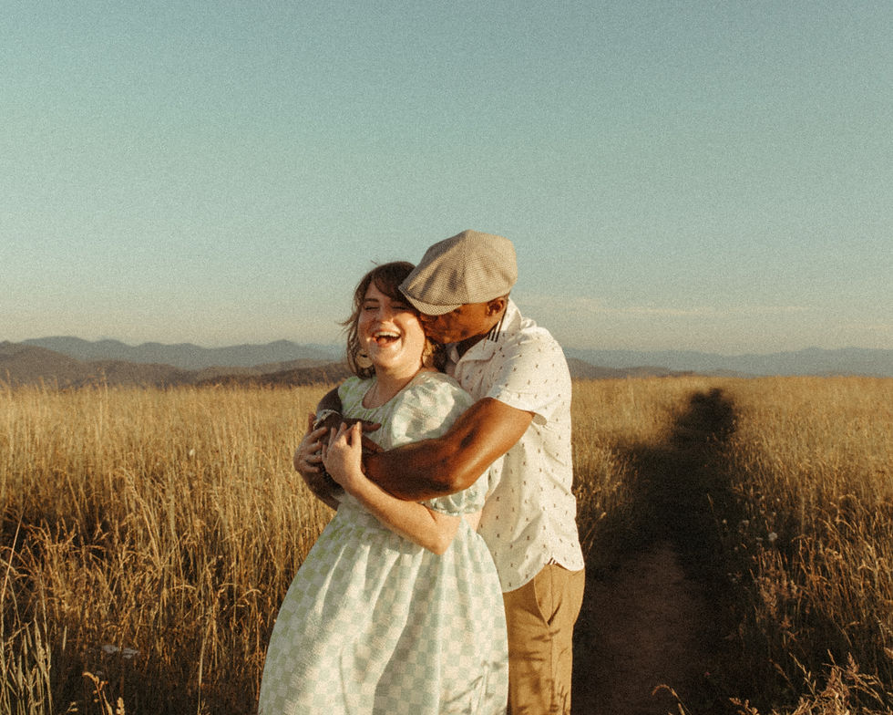 Kevin holding Kate close in a golden field, both laughing, dressed in casual warm-weather clothing with an open sky and dry grass in the background — a candid moment of joy and connection between two partners.