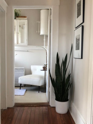 A quiet nook with a white chaise lounge, plant stand, and soft draped fabric, seen through an open doorway