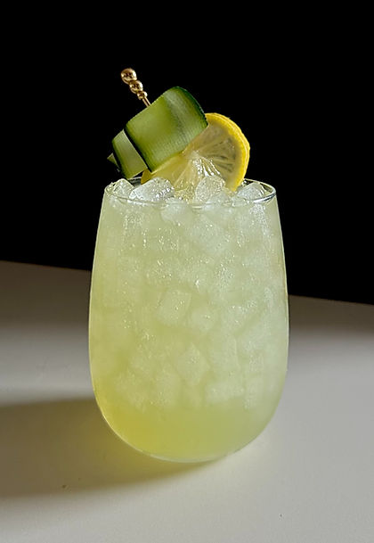 A pale green cucumber celery mocktail served over crushed ice in a stemless glass, garnished with a long cucumber ribbon and a lemon wheel.