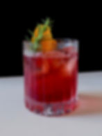 Deep red Americano cocktail in a cut crystal rocks glass with two large ice cubes, an orange twist, and a fresh rosemary sprig, white surface, black background.