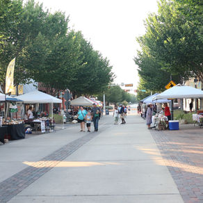 A downtown street lined with vendor tents on both sides, featuring tables with goods and shoppers walking along the center under tall, leafy trees during an outdoor market event.