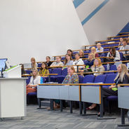 Presenter speaking at the front of a lecture theatre while attendees listen.