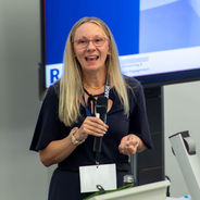 Presenter delivering a conference session while holding a microphone beside presentation equipment.