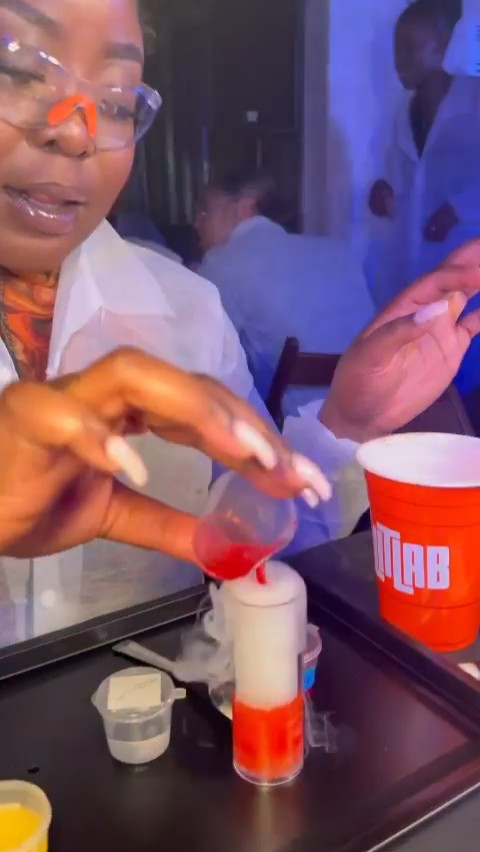 a woman in goggles and a lab coat pours liquid into a smoky shot cup at the lit lab london