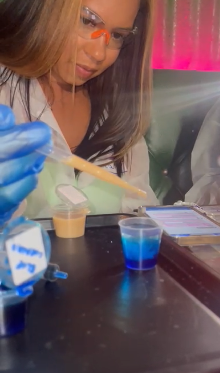a woman in gloves goggles and a lab coat uses a pipette to drop liquid into a shot cup with blue layered liquids at the lit lab london