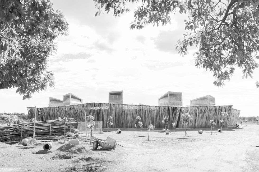 Black-and-white view of a modern building under construction, framed by trees, with a wheelbarrow, pipes, and small plants in front.