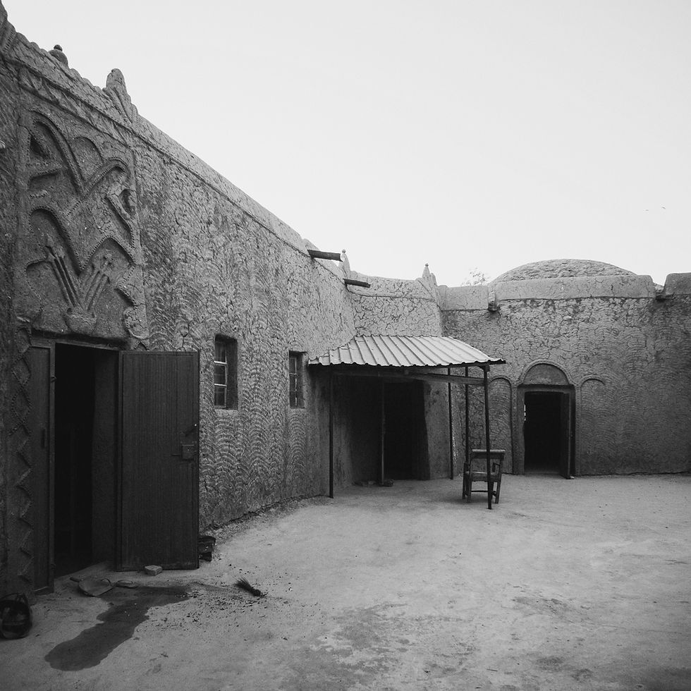 Black-and-white courtyard of an earthen building with open doorways, a corrugated awning, and a stark, empty, quiet mood.