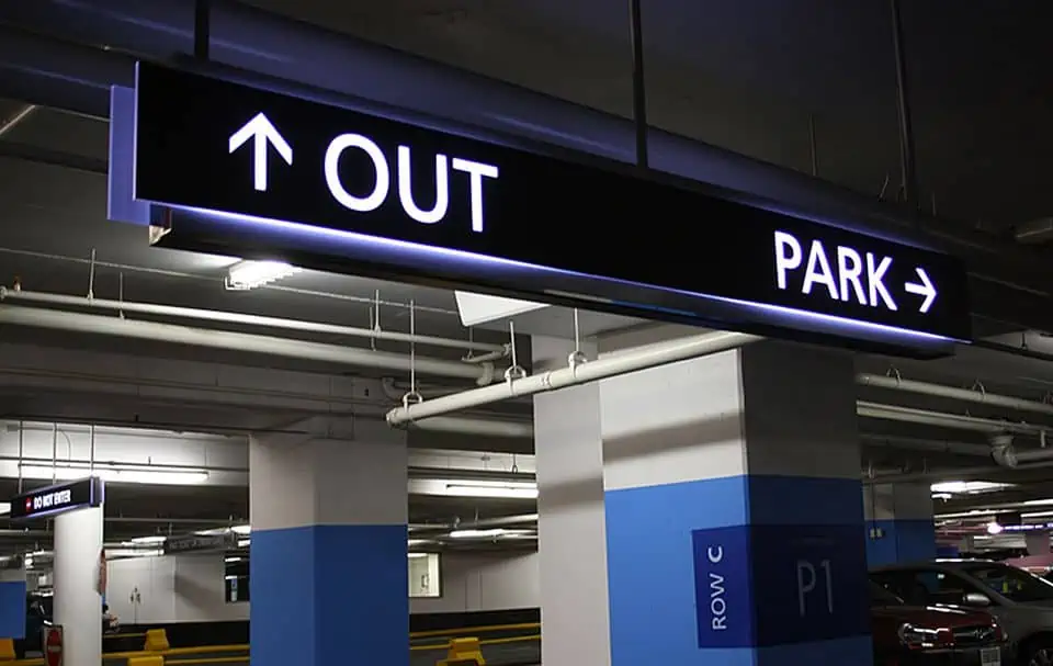A hanging Parking Lot Sign that is backlit. It is directing people to the exit and parking