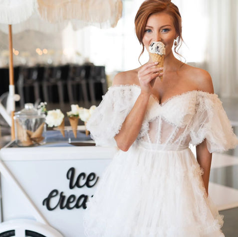 Woman in wedding dress eats ice cream next to "Ice Cream" cart.