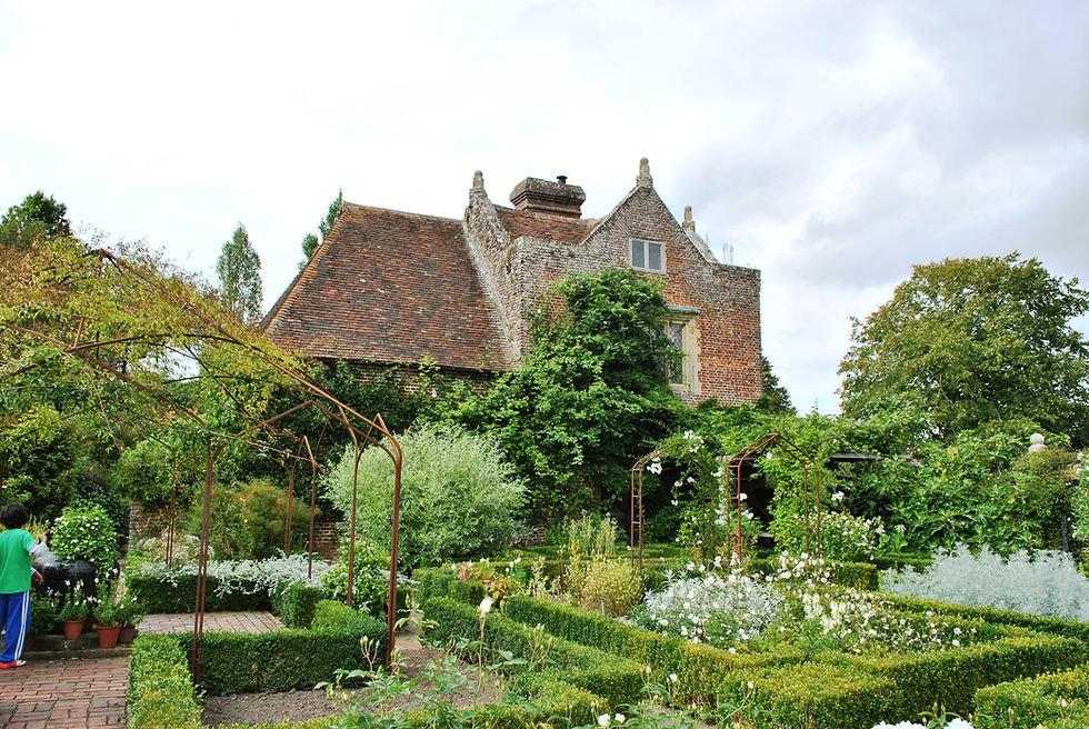 Historic brick house with a lush garden in a Sissinghurst Castle Garden (Kent). A child waters plants under a metal arbor. Greenery and flowers create a serene, natural setting.