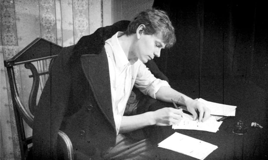 Young man poet Sergei Yesenin writing at a table, wearing a coat over shoulders. Papers and ink bottle are on the dark table. Focused expression, vintage setting.