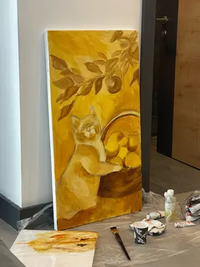An oil painting made by artist Yana Evans in its early stages rests against a wall, showing a rough sketch of a cat holding a basket of lemons beneath leafy branches, all in warm yellow and brown tones. Art supplies including paint tubes, a brush, and a mixing board lie on a plastic-covered floor nearby, capturing the primal layer and first brushstrokes of the creative process.
