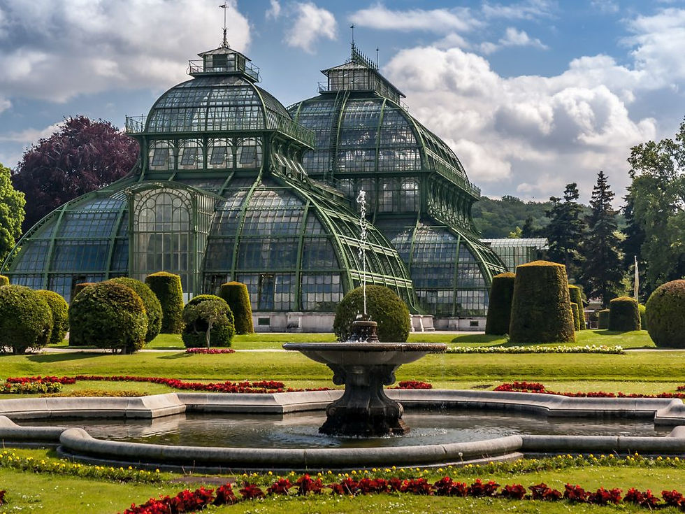 Grand greenhouse with ornate glass domes in a manicured garden in a Schönbrunn Palace gardens. Central fountain and vibrant flowerbeds create a serene atmosphere.