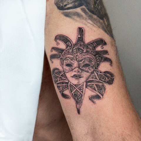 Tatuaje en negro y gris de una máscara ornamental de bufón de estilo veneciano con detalles de filigrana, tatuada en la parte superior del brazo por Yana Evans (evans ink).