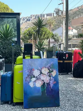 A collection of colorful suitcases, including bright yellow, blue, red, and black, is gathered outdoors on a stone-paved surface, with a scenic backdrop of palm trees, power lines, and hillside buildings. In front of the luggage stands an unfinished oil painting of white and pink flowers with purple blossoms in a green vase, created by artist Yana Evans. The vivid painting rests against a yellow suitcase, capturing a moment of artistic travel preparation.