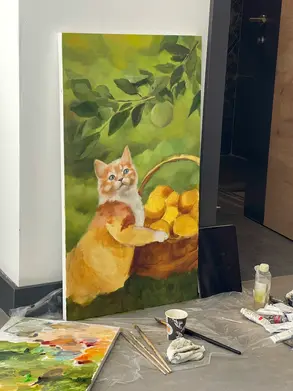 An in-progress oil painting by artist Yana Evans resting against a wall in a hotel room, showing a ginger and white cat with a basket of yellow lemons under green leaves. The painting is on the floor near a bathroom door, surrounded by paintbrushes, a palette, a paper cup, and oil paint tubes, capturing a cozy, creative moment.
