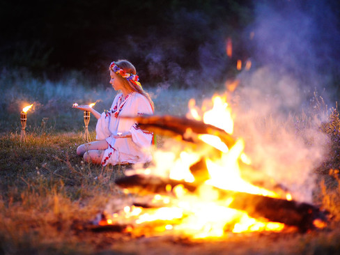 Young woman in traditional Slavic dress kneeling by a midsummer bonfire at night, part of a Kupala Night ritual with floral wreaths and torchlight ceremony.