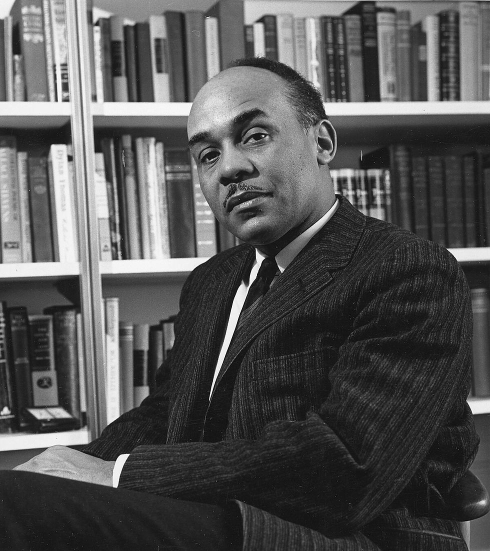 Ralph Ellison, con un traje oscuro de rayas, está sentado frente a una estantería llena de libros. Mira con serenidad a la cámara. Fotografía en blanco y negro.