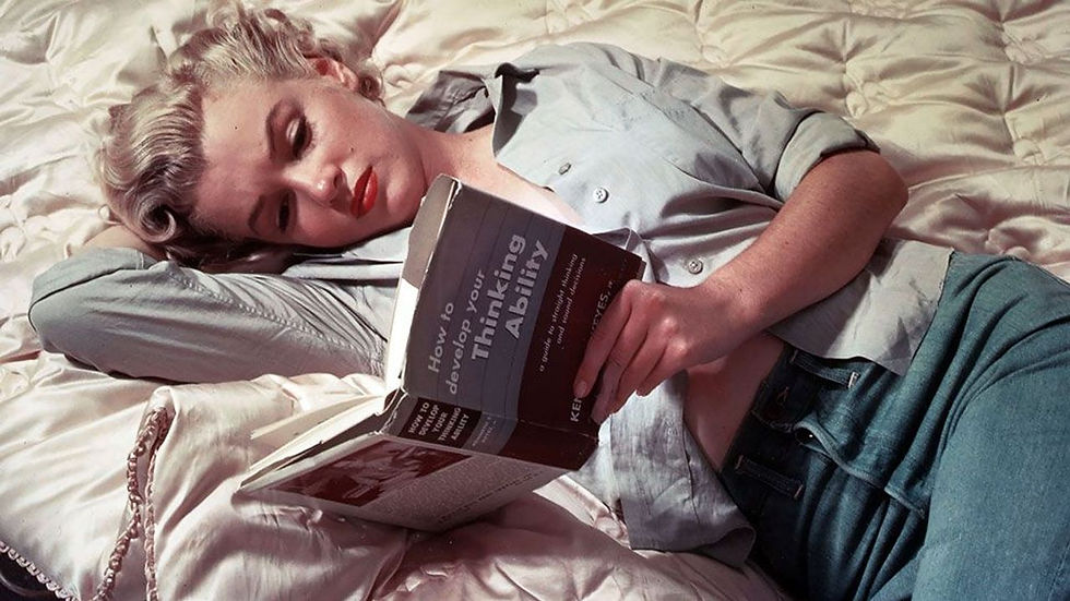 Marilyn Monroe, con el cabello rubio, labios rojos y una camisa celeste, yace sobre una cama de satén leyendo Cómo desarrollar tu capacidad de pensar.