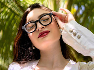 Close-up of a woman in black glasses and a white blazer adjusting them, with sunlit palm leaves blurred behind her.