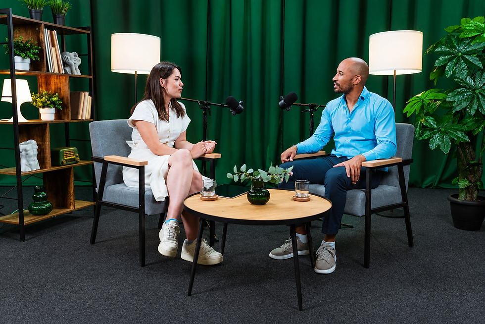 Studio photography of a man and woman in a podcast interview. They are sitting in comfortable chairs with microphones between them, set against a dark green backdrop.