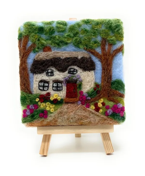 View of a finished 2d thatched cottage needle with trees and flowers around, standing on a wooden easel.