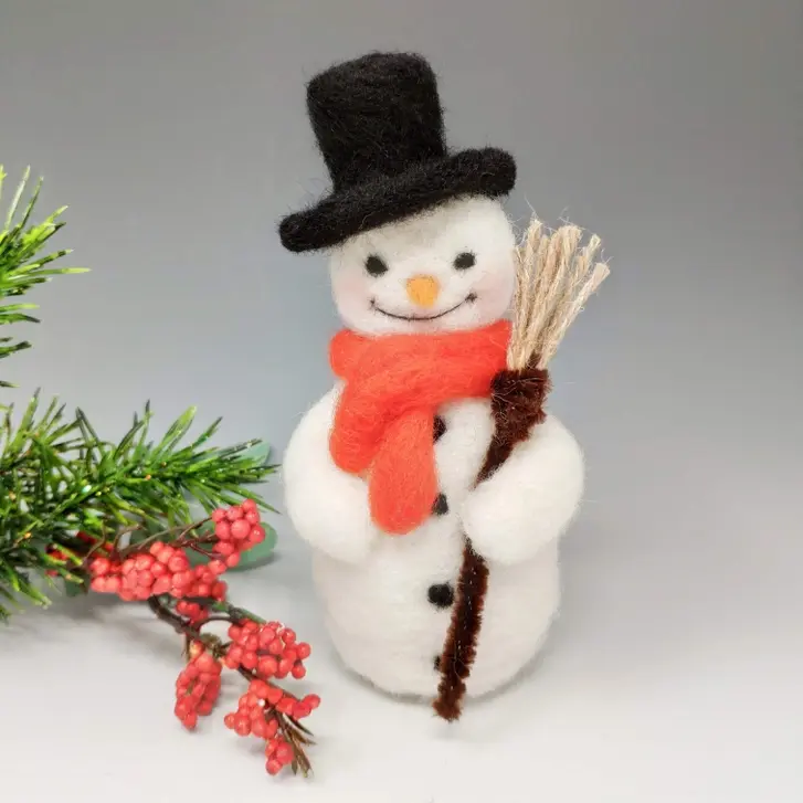 needle felted snowman with an orange scard, black hat holding a broom beside some green foliage and red berries