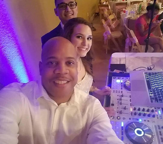 DJ Undaground smiling with a newlywed couple at a wedding reception, standing behind a professional DJ setup