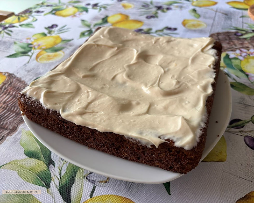 Gâteau aux carottes sans gluten carré, recouvert d'un glaçage crémeux couleur vanille, sur une assiette blanche avec une nappe à motifs de citrons et d'olives en arrière-plan.
