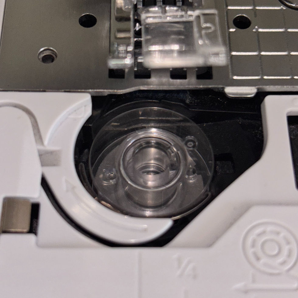 Close-up of a sewing machine bobbin case with the correct bobbin, in a metal and white housing. Visible grid pattern and partial view of presser foot above.