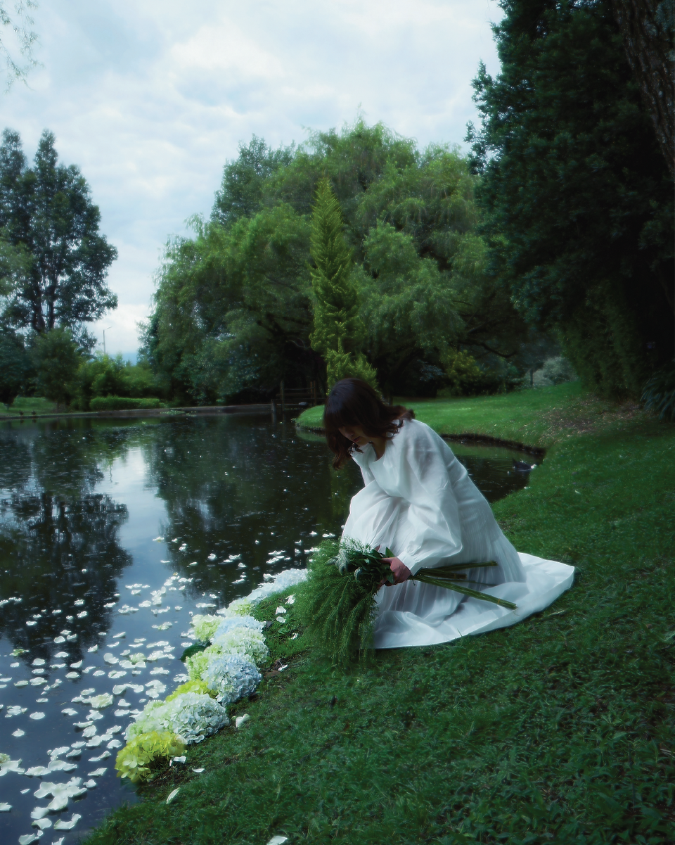 A woman dressed in flowing white delicately arranges green and white hydrangeas by a reflective pond, surrounded by lush trees. The floating petals and serene atmosphere capture the mystical essence of nature, evoking the enchantment of St. Patrick’s season.