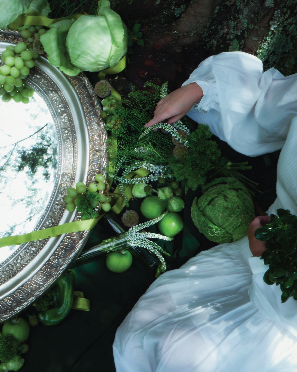 A woman in a flowing white dress arranges green flowers and foliage beside an ornate silver mirror, surrounded by fresh apples, grapes, and cabbages. The interplay of reflections and natural elements evokes the mysticism of St. Patrick’s season, celebrating the harmony between florals and the earth’s bounty.