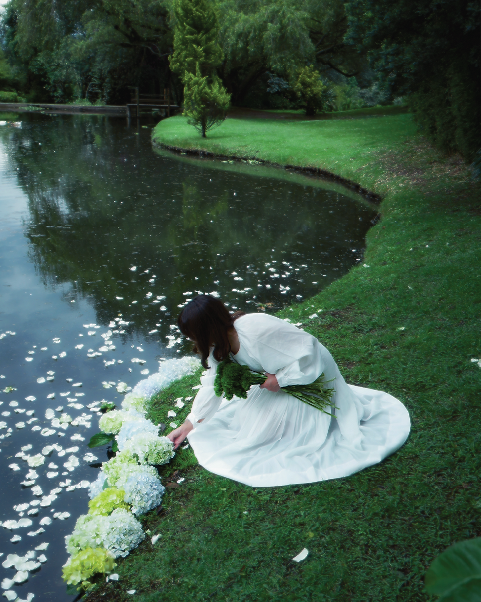 A woman in a flowing white dress kneels by a peaceful pond, arranging delicate green and white hydrangeas along the water’s edge. White petals float on the surface, blending with the tranquil landscape, evoking the magic and renewal of St. Patrick’s season through nature’s beauty.
