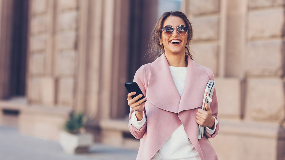 Woman in a pink coat, smiling, holds a phone and a folder. Sunglasses and urban background suggest a cheerful, sunny day.
