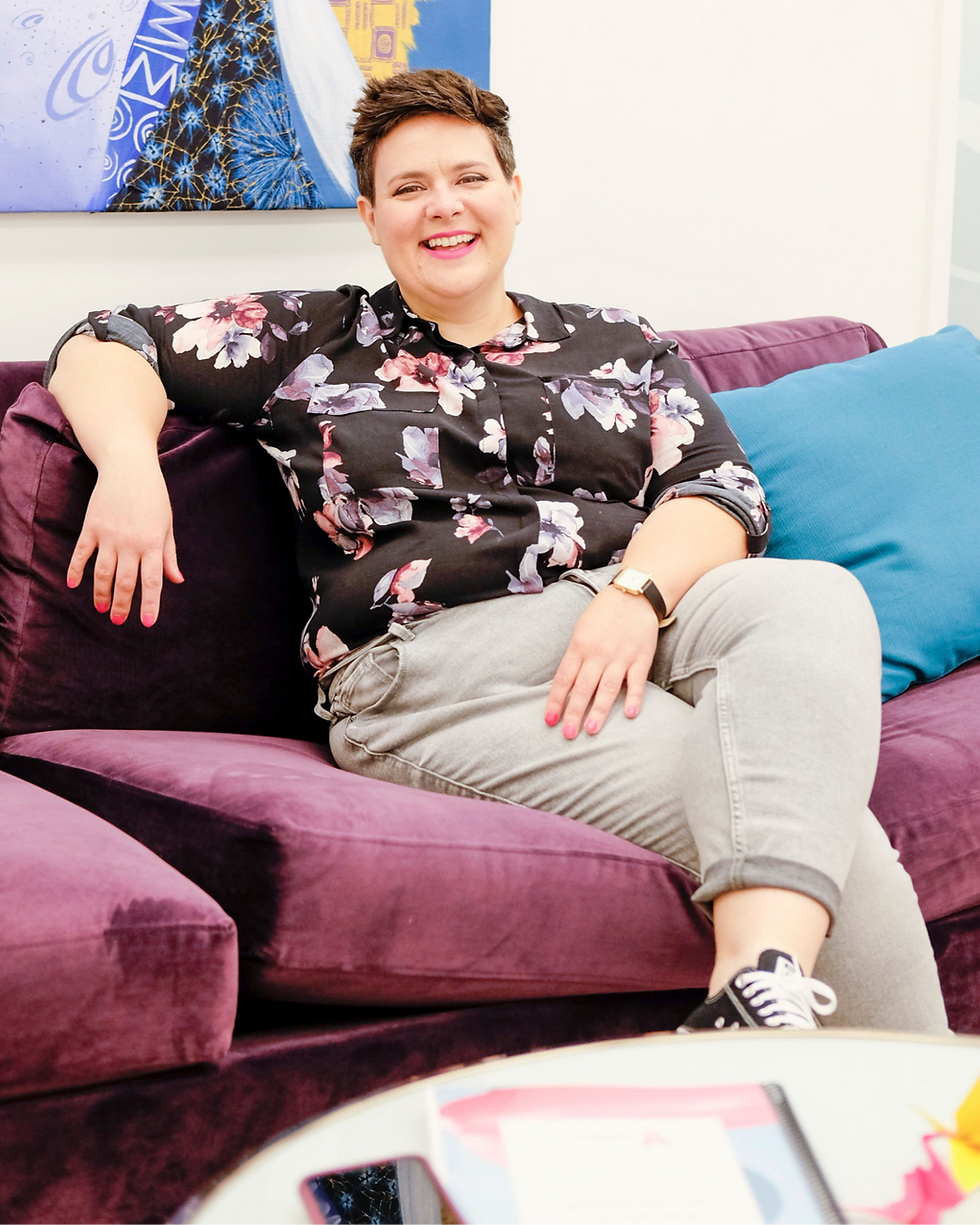 Smiling person in a floral shirt sits on a purple couch with blue pillows. Colorful abstract art in the background. Relaxed and cheerful mood.