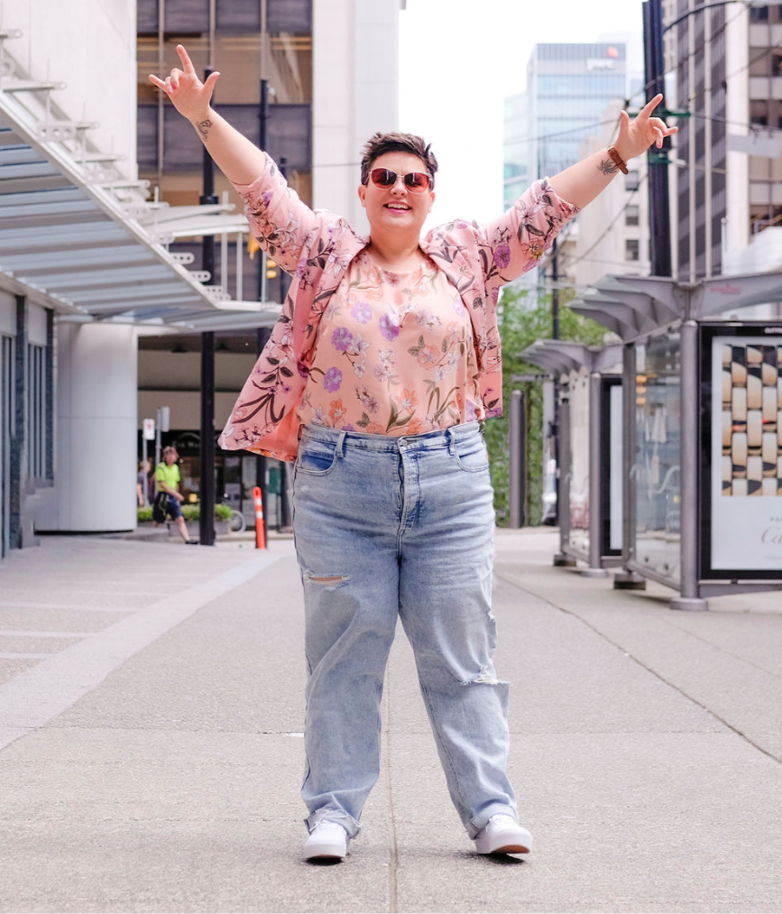 A person in floral pink attire and ripped jeans joyfully raises arms in a city street. Red truck, Cartier ad, and buildings in background.