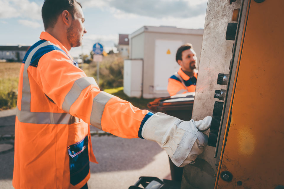 two males, facing each other, on the back of a garbage truck wearing reflective orange vests.