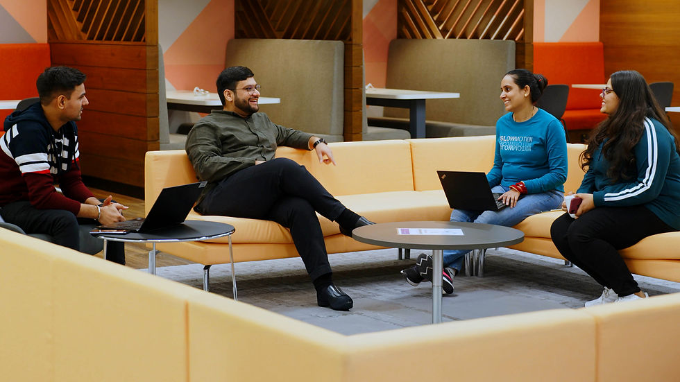 Four people sit and chat on orange sofas with laptops in a modern, cozy lounge. One wears a blue top reading "SLOWMOTION."