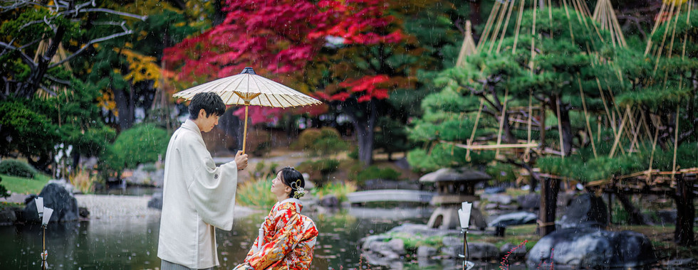 Bride in colorful Iro-Uchikake wedding kimono sitting beside a traditional Japanese garden with autumn foliage.