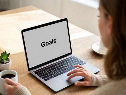 Person viewing laptop screen displaying "Goals" at a wooden table, holding a coffee cup. Small green plant in the background.
