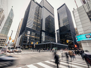Busy city intersection with blurred pedestrians crossing. Tall glass skyscrapers surround, reflecting urban life, with traffic lights and cars.