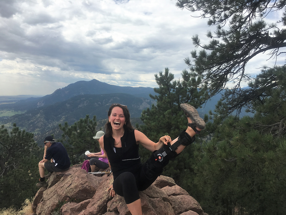 A young woman with brown hair, sunglasses on top of her head, and covered in all-black workout gear is laughing as she sits on a mountain peak and holds up her leg, which is in a post-surgery immobilizer. There are two other, older people in the background also on the peak. Further in the background, green, tree-covered mountains pop up, with clouds sweeping across the sky.