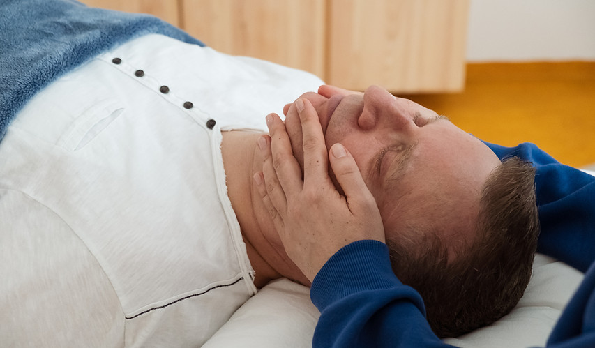 Mann liegt entspannt auf einem Massagetisch, während eine cranio-sacrale Behandlung am Kiefer durchgeführt wird.