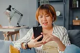 young-asian-lady-using-smart-phone-video-call-talk-with-family-sofa-living-room-house-scal