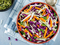 Colourful coleslaw salad with purple cabbage, white cabbage, carrots, and seeds in a bowl on a blue-checkered cloth, with a spoon and seeds scattered nearby.