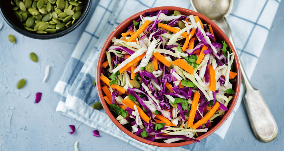 Colourful coleslaw salad with purple cabbage, white cabbage, carrots, and seeds in a bowl on a blue-checkered cloth, with a spoon and seeds scattered nearby.