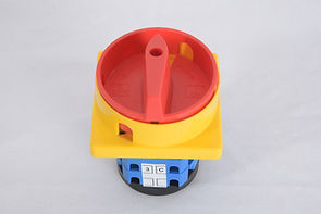The image shows a rotary switch with a red rotary knob and a handle, mounted on a yellow square base. Below the base, there's a blue and black component with white labels that likely indicate electrical connections or settings. This type of switch is commonly used for controlling electrical circuits and can be found in industrial or machinery applications.