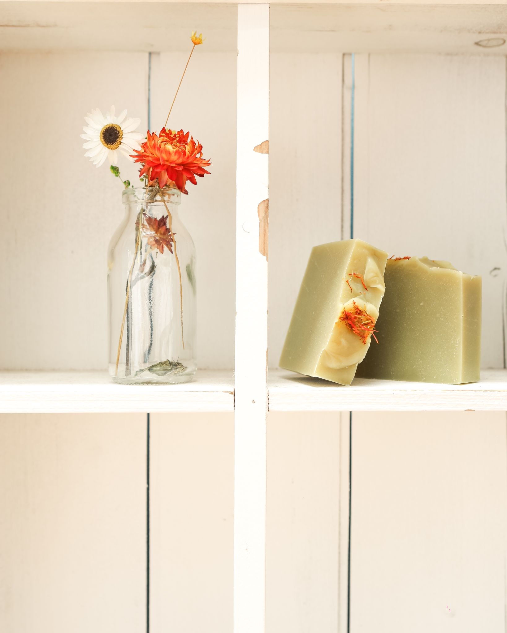 green and orange soap bars leaning against each other in a cupboard with a glass bottle vase with orange and white dried flowers