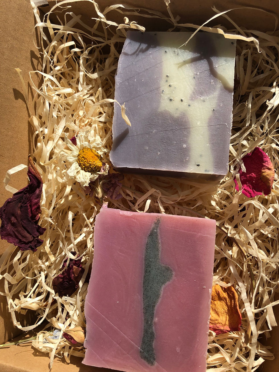 Thumbnail: A natural gift box containing woodwool infused with dried botanicals and a pink soap with a grey strip centrally and a purple soap with a white strip containing poppy seeds centrally.