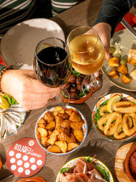 Zwei Personen stoßen mit Rot- und Weißwein über einem gedeckten Tapas-Tisch an.